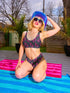 Two-piece bikini set featuring a bold pastel goth design with neon pink skulls wearing green bows, surrounded by multicolored stars on a deep black background. The set includes a plunging V-neck bikini top with wide straps and high-waisted bottoms for a flattering retro fit. Made of stretchy, soft material with a smooth black lining. This skull print swimsuit is ideal for alternative, edgy summer style with a playful punk twist.